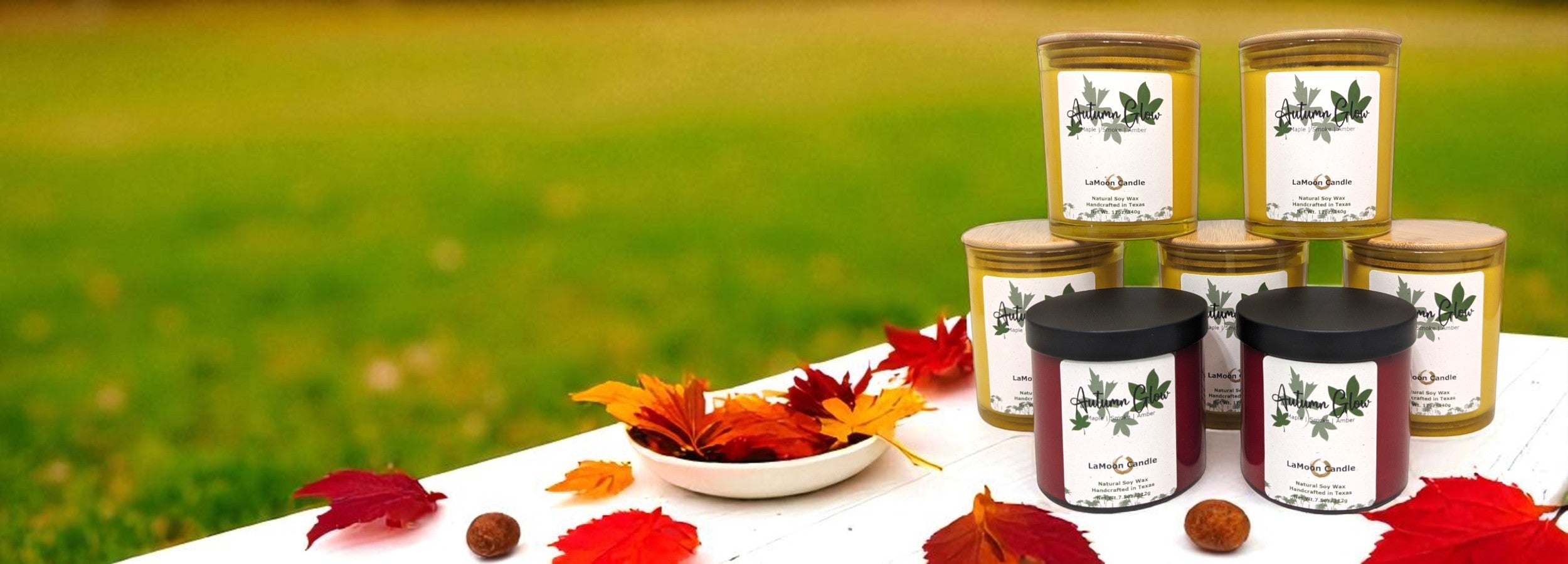 Fall Collection Candles with decorative labels on a table with autumn leaves against a grassy background. 