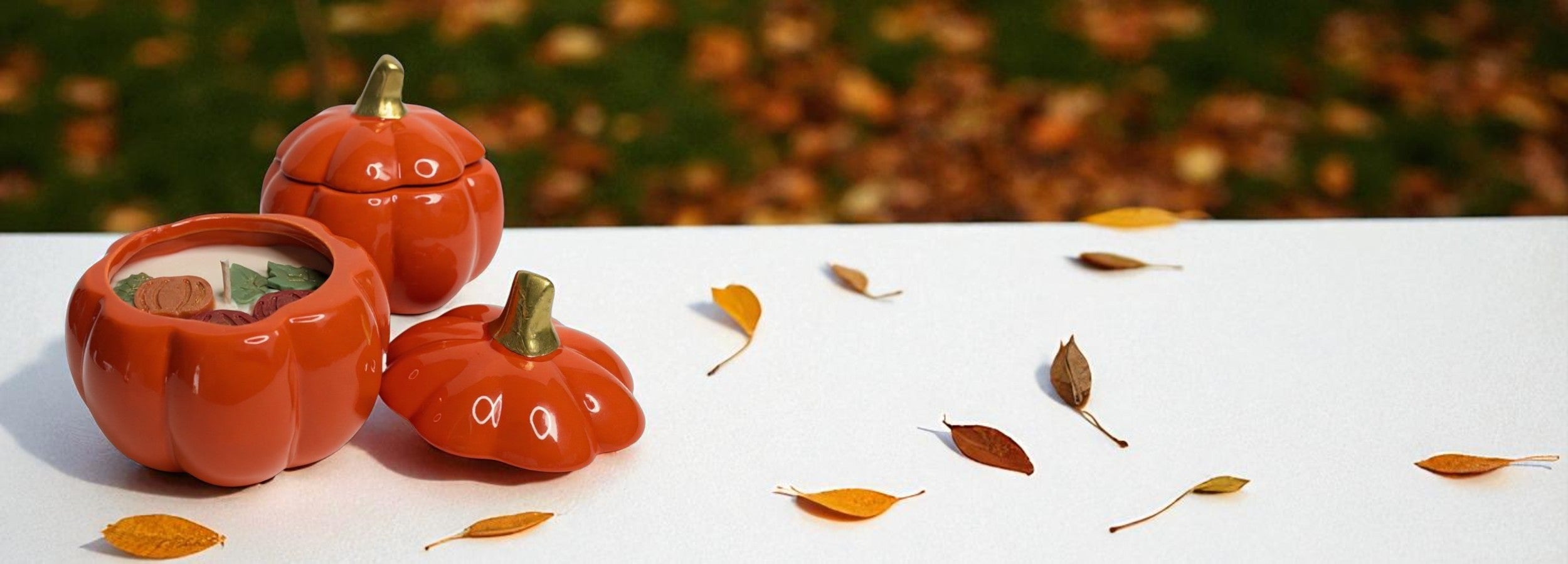 Ceramic pumpkin-shaped containers with soy wax candles on a white surface with autumn leaves.