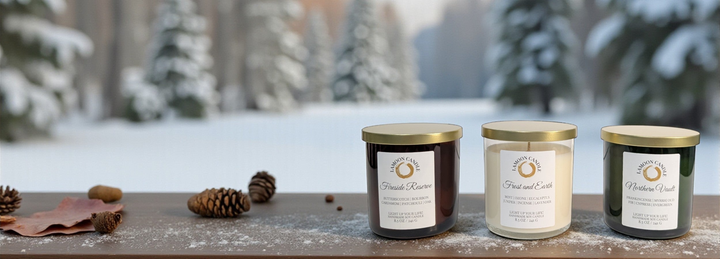 Three LaMoon Candle's candles with labels on a wooden surface in front of a snowy forest background. There are three scent; Fireside Reserve, Frost and Earth, and Northern Vault.