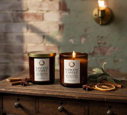 Two LaMoon Candle scented soy candles in amber glass jars with white labels and gold lids, lit and displayed on a rustic wooden table with cinnamon sticks, star anise, and dried orange slices against a brick and painted wall background.