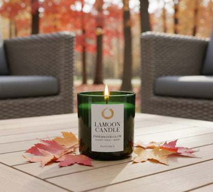 A green glass jar candle with a white label reading 'LAMOON CANDLE' and 'EMBERWOOD GLOW' is lit on a light wood table outdoors. Fallen autumn leaves are scattered around the candle, with blurred patio chairs and trees in the background.