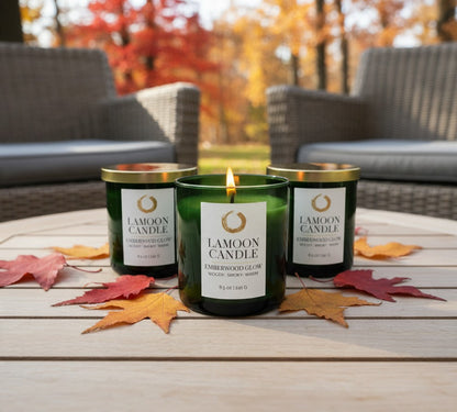 Three LaMoon Candle jars arranged on a light wood table outdoors, with autumn leaves scattered around and wicker patio furniture blurred in the background. The center candle is lit and in front, with two unlit candles behind it.